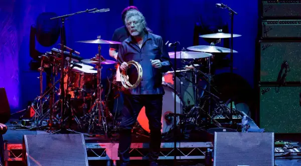 Robert Plant z zespołu zeppy zep, trzymający tamburyn na scenie, w tle perkusja i wzmacniacze.