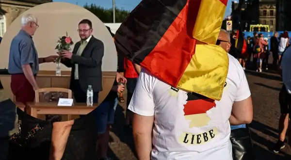 Mężczyzna w koszulce z mapą Niemiec i napisem "Liebe" niesie flagę Niemiec. W tle widać ludzi i budynki.