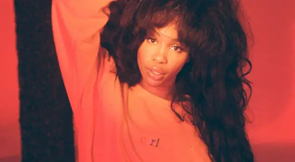 SZA w pomarańczowej bluzie z napisem "crl", z ręką we włosach na tle czerwonym.