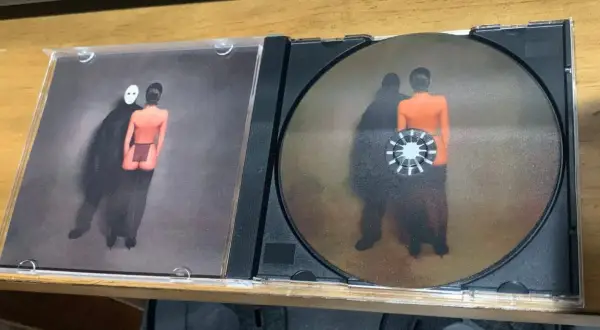 Otwarta płyta CD z okładką przedstawiającą postać w masce i nagą postać. Na płycie widnieje podobny obraz, jakby te postacie były jak sępy, ye.