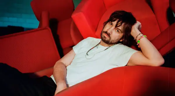 Kevin Parker z Tame Impala Polska odpoczywa na czerwonej kanapie, otoczony neonowym światłem.