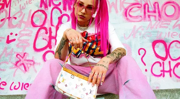 Młoda Leosi w różowych włosach i spodniach, z torebką w ręku, na tle graffiti.