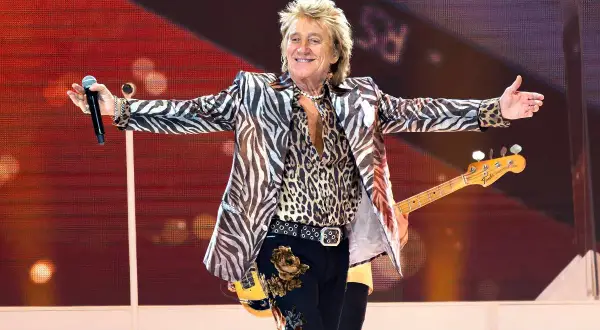 Rod Stewart w panterce i marynarce w zebrę, z gitarą basową, na scenie, jakby to był największy koncert na świecie.