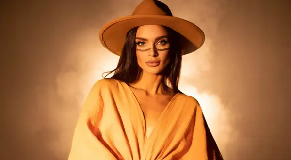 DJ Korolova w kapeluszu i pomarańczowej sukience, z ozdobnym makijażem na twarzy, emanuje tajemniczością i stylem.
