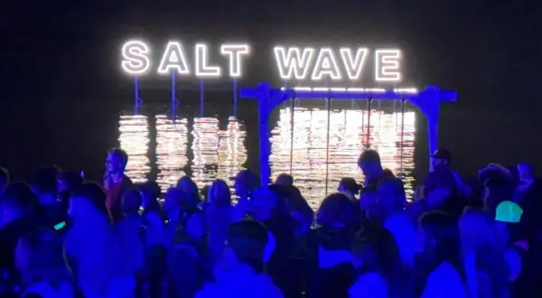 Nocny tłum na festiwalu Salt Wave w Jastarni. Neonowy napis odbija się w wodzie.