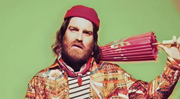Chet Faker w czerwonej czapce i kolorowej kurtce trzyma różowy parasol.