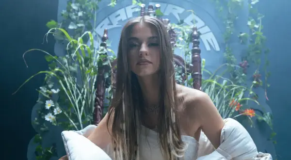 Młoda kobieta w białej sukni, otoczona bujną zielenią, siedzi na ozdobnym tronie. W tle widnieje napis "Bambi Rap".