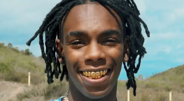 YNW Melly z dredami i złotymi zębami uśmiecha się na tle krajobrazu.