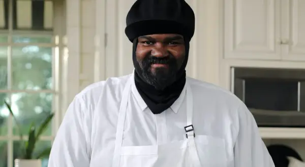 Gregory Porter w białej koszuli i czarnej czapce, z brodą, uśmiecha się w kuchni.