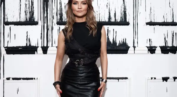 Carla Roca w czarnym, skórzanym stroju pozuje na tle ściany z czarnymi, spływającymi wzorami.