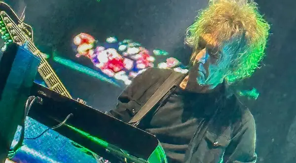 Perry Bamonte z burzą blond włosów, grający na gitarze i syntezatorze podczas koncertu, otoczony kolorowymi światłami.