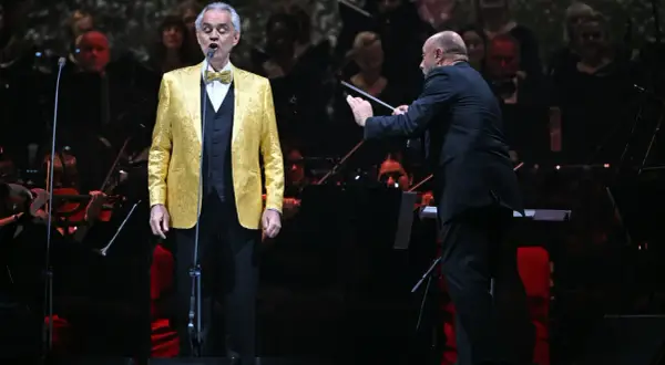 Andrea Bocelli w złotej marynarce śpiewa na koncercie w Warszawie, dyrygent prowadzi orkiestrę.