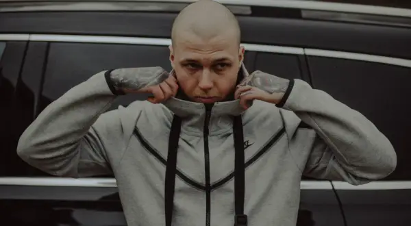 Szpaku golem, z tatuażami na rękach, poprawia kołnierz szarej bluzy Nike, opierając się o czarny samochód.