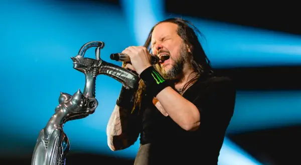 Jonathan Davis z **Korn Polska** śpiewa z pasją do mikrofonu, trzymając go na tle niebieskiego światła.