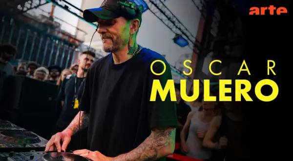 Oscar Mulero, DJ z tatuażami, miksuje muzykę na żywo, podczas gdy tłum ludzi bawi się w tle. Arte.