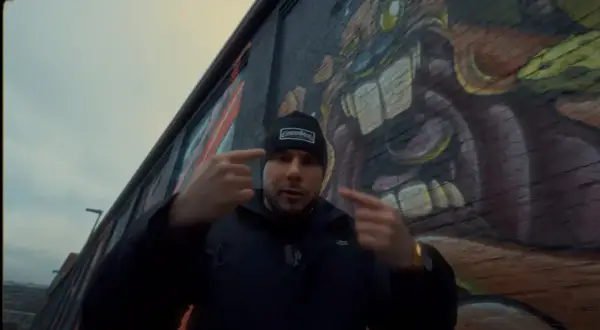 Mężczyzna w czapce z napisem "GermanStreets" wskazuje palcami na siebie, stojąc przed ścianą z graffiti. Może to być zapowiedź bonusa rpk.