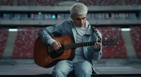 Artysta z blond włosami gra na gitarze w pustym stadionie. Zapowiada swój nowy album.