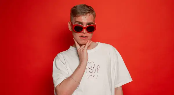 Młody mężczyzna w czerwonych okularach i białej koszulce z rysunkiem twarzy, jakby szykował się do występu koza rap.