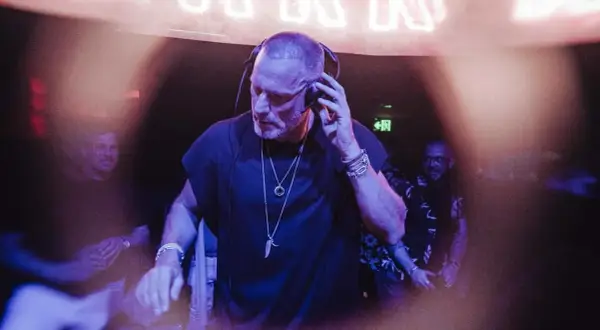 Chriss Jaxx w akcji, miksuje muzykę w klubie. W tle neonowy napis i ludzie bawiący się w rytm jego setu.