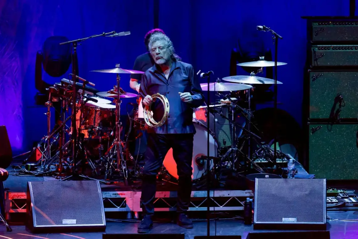 Robert Plant z zespołu zeppy zep, trzymający tamburyn na scenie, w tle perkusja i wzmacniacze.