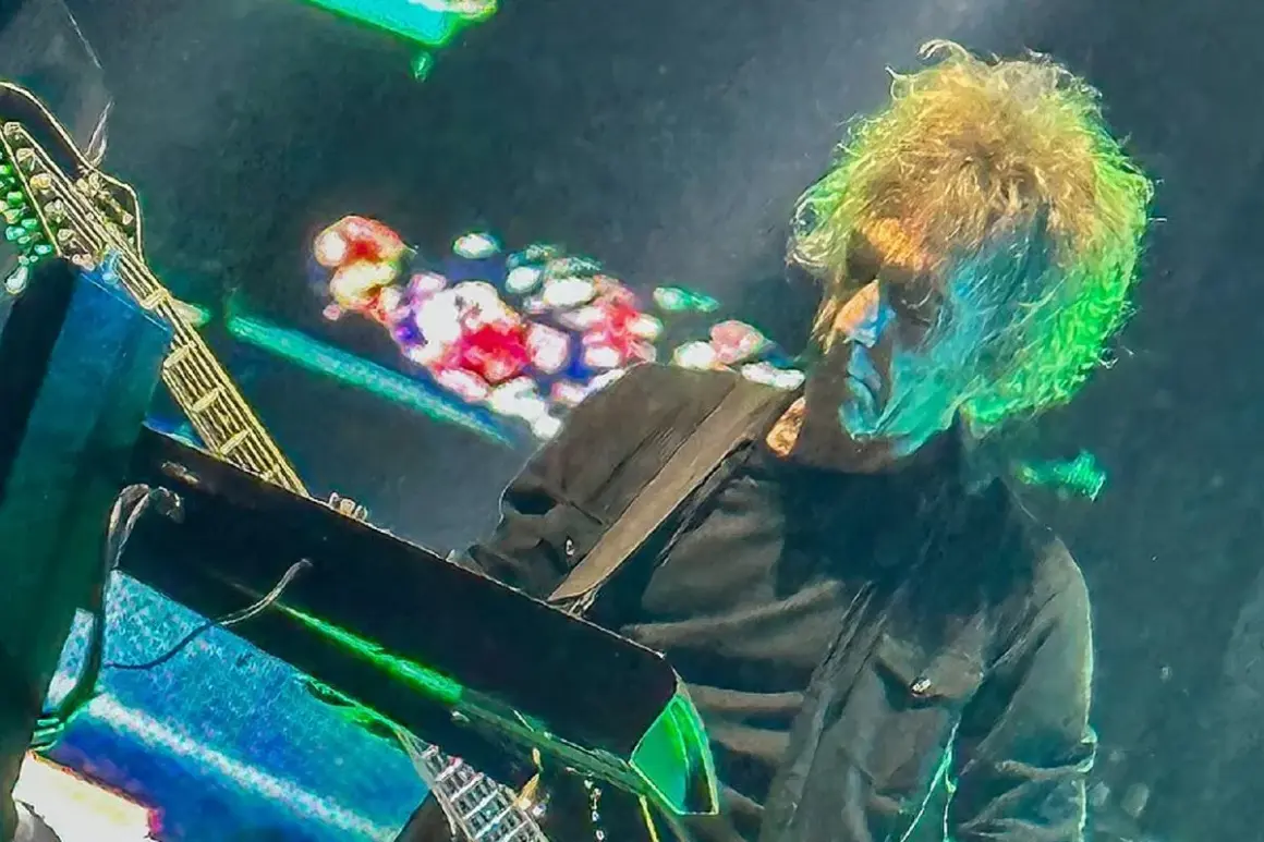 Perry Bamonte z burzą blond włosów, grający na gitarze i syntezatorze podczas koncertu, otoczony kolorowymi światłami.