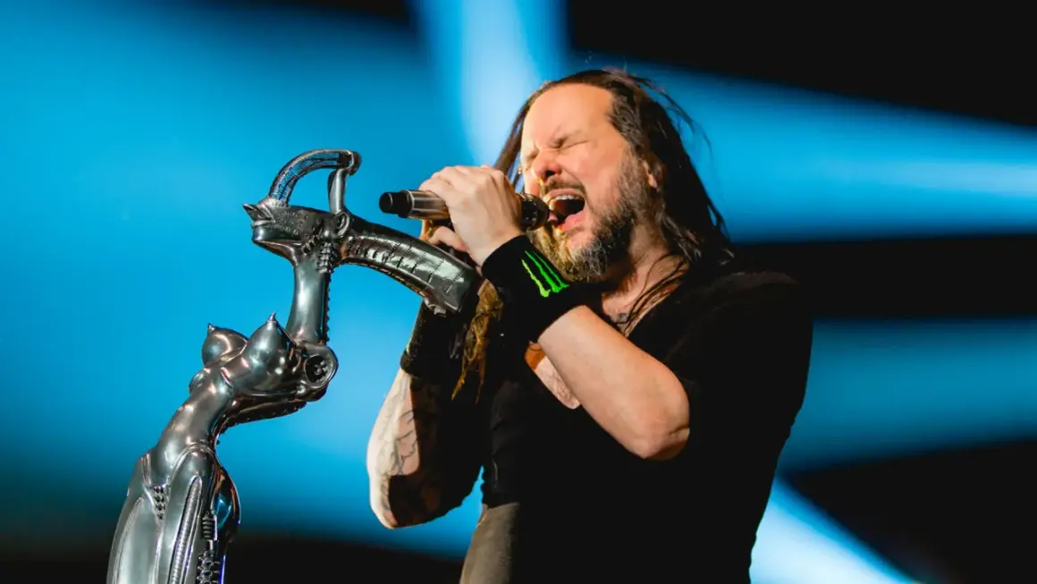 Jonathan Davis z **Korn Polska** śpiewa z pasją do mikrofonu, trzymając go na tle niebieskiego światła.