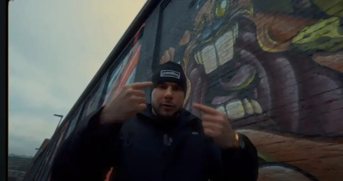 Mężczyzna w czapce z napisem "GermanStreets" wskazuje palcami na siebie, stojąc przed ścianą z graffiti. Może to być zapowiedź bonusa rpk.