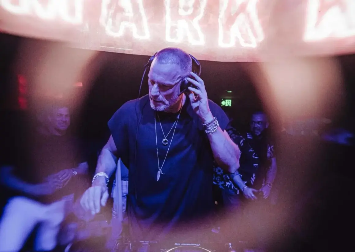 Chriss Jaxx w akcji, miksuje muzykę w klubie. W tle neonowy napis i ludzie bawiący się w rytm jego setu.