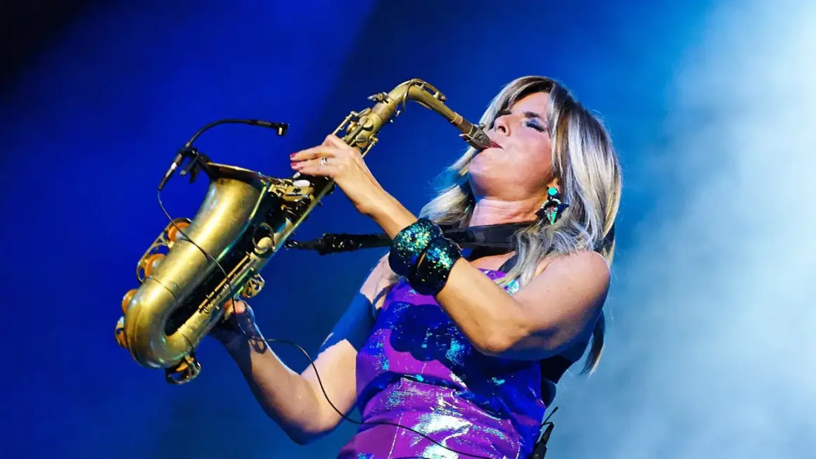 Candy Dulfer gra na saksofonie, jej blond włosy opadają na fioletową, błyszczącą sukienkę.