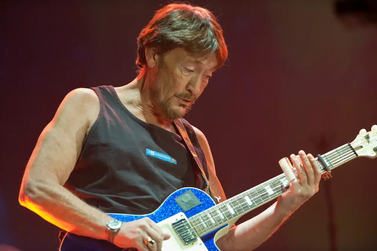 Chris Rea, w czarnej koszulce bez rękaw&oacute;w, gra na błyszczącej, niebieskiej gitarze elektrycznej.