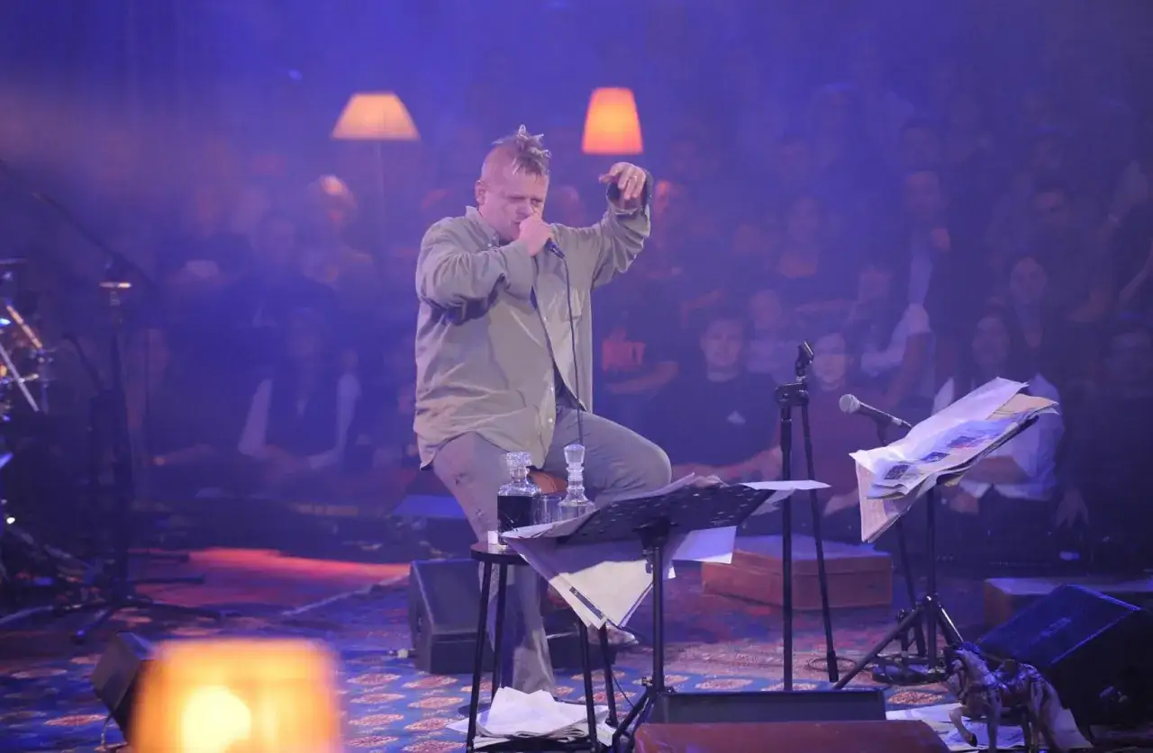 Kazik Staszewski podczas MTV Unplugged Polska, siedząc na stołku, śpiewa do mikrofonu. W tle widać publiczność i scenografię z lampami.