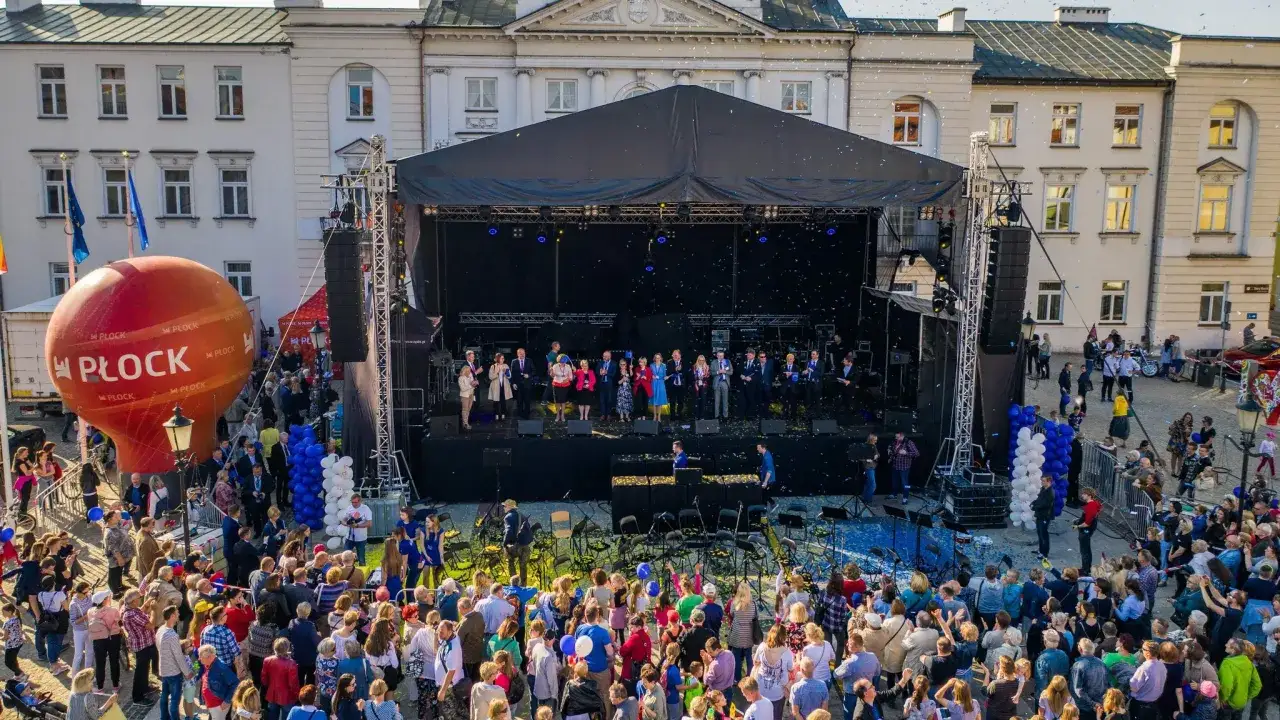 Wielki czerwony balon z napisem Płock unosi się nad placem, gdzie trwa festiwal. Na scenie występują artyści, a tłum widzów świętuje.