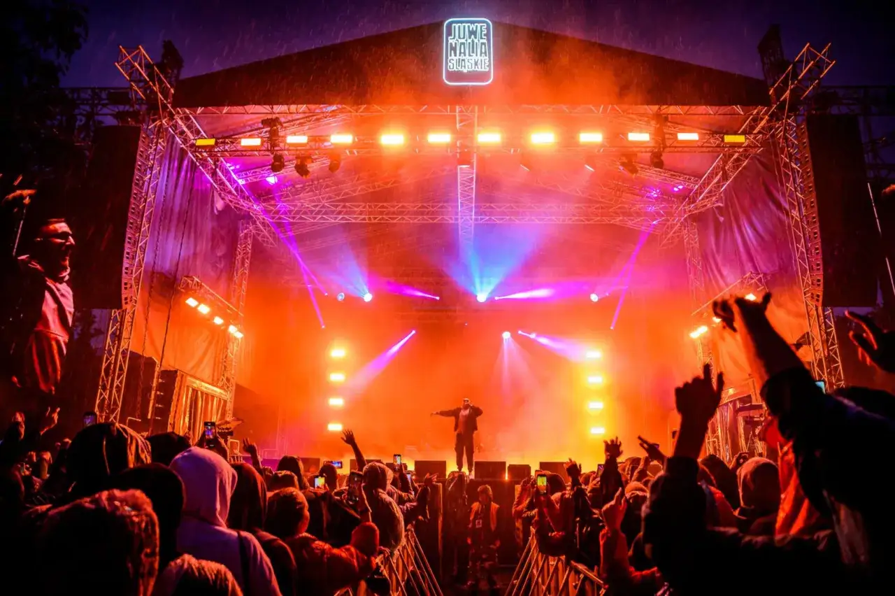 Koncert na Juwenaliach Katowice. Tłum szaleje pod sceną oświetloną pomarańczowymi i fioletowymi światłami, w deszczu.