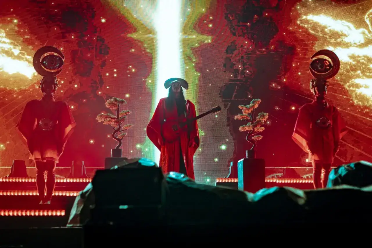 Na scenie, w blasku czerwieni i złota, artysta w stroju przypominającym empire of the sun gra na gitarze. Po bokach stoją dwie postacie w czerwonych szatach z ozdobami na głowach.