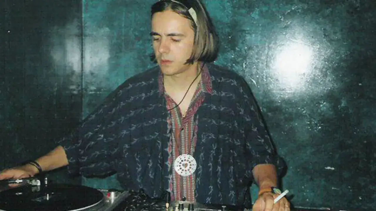 DJ z lat 90. w słuchawkach, tworzący muzykę techno. W tle mroczna, klubowa atmosfera.