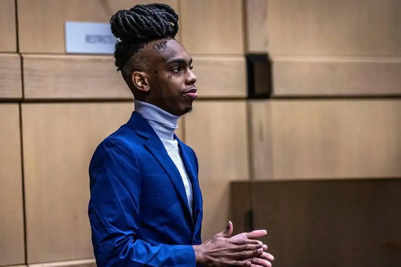 YNW Melly w niebieskiej marynarce i szarym golfie, z charakterystyczną fryzurą i tatuażami na twarzy, stoi w pomieszczeniu z drewnianymi ścianami.