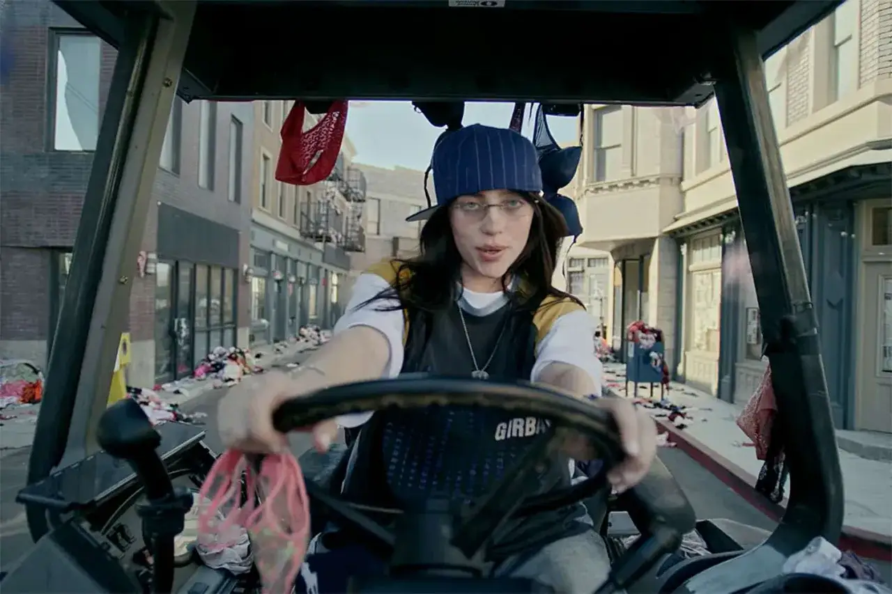 Billie Eilish, jak brat, prowadzi w&oacute;zek widłowy przez ulicę zasypaną ubraniami.