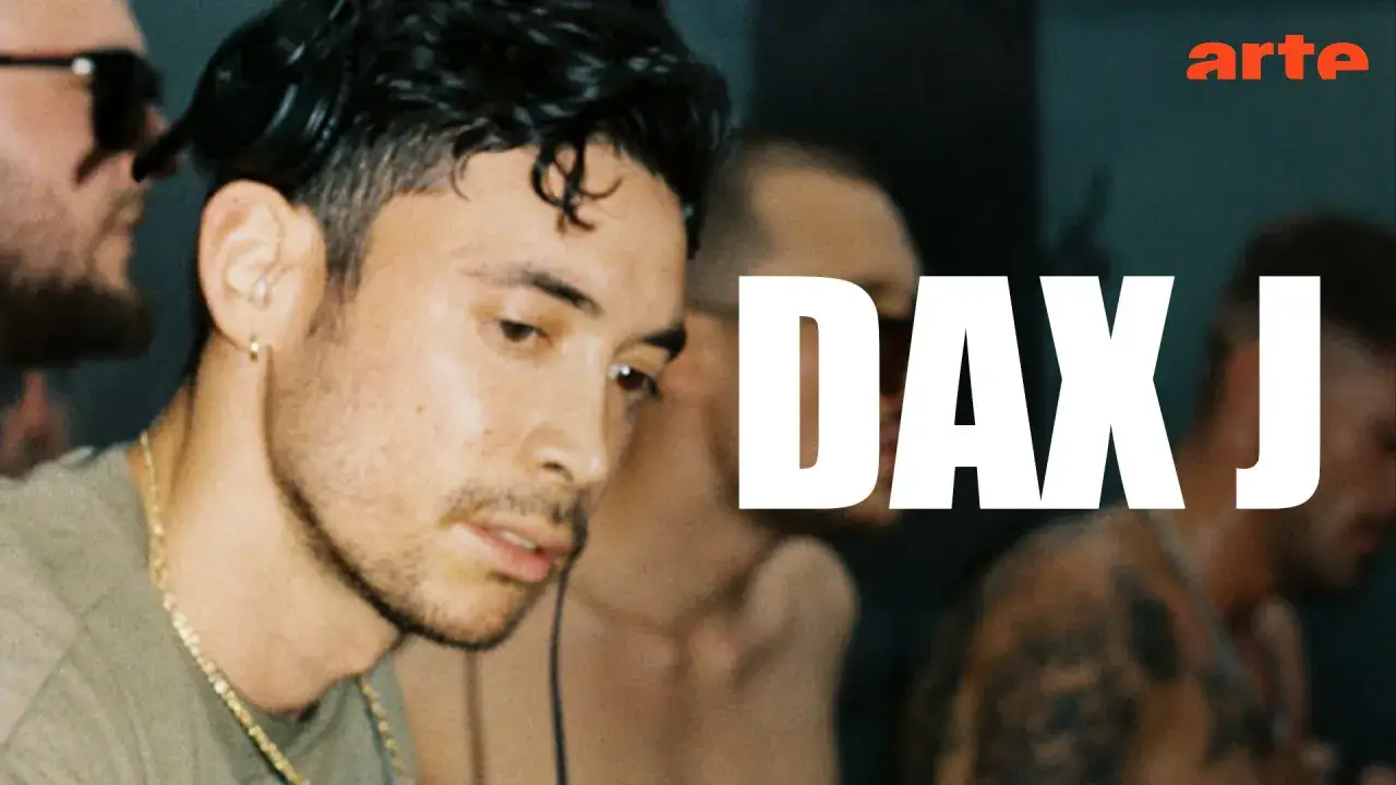 Dax J, artysta z charakterystycznym kolczykiem w uchu i złotym łańcuchem, w otoczeniu innych osób.
