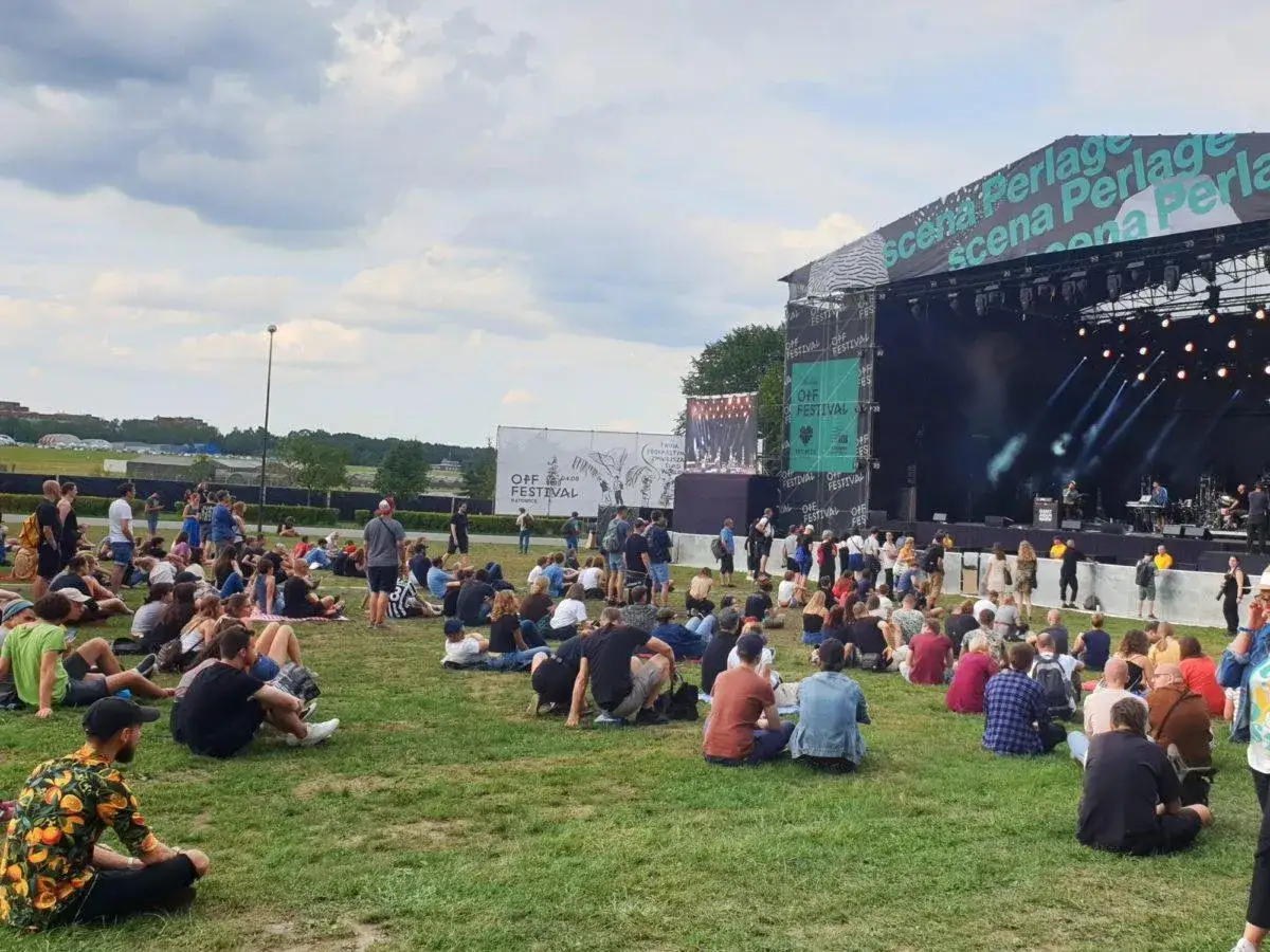 Tłum ludzi siedzi na trawie przed sceną OFF Festival, ciesząc się muzyką.
