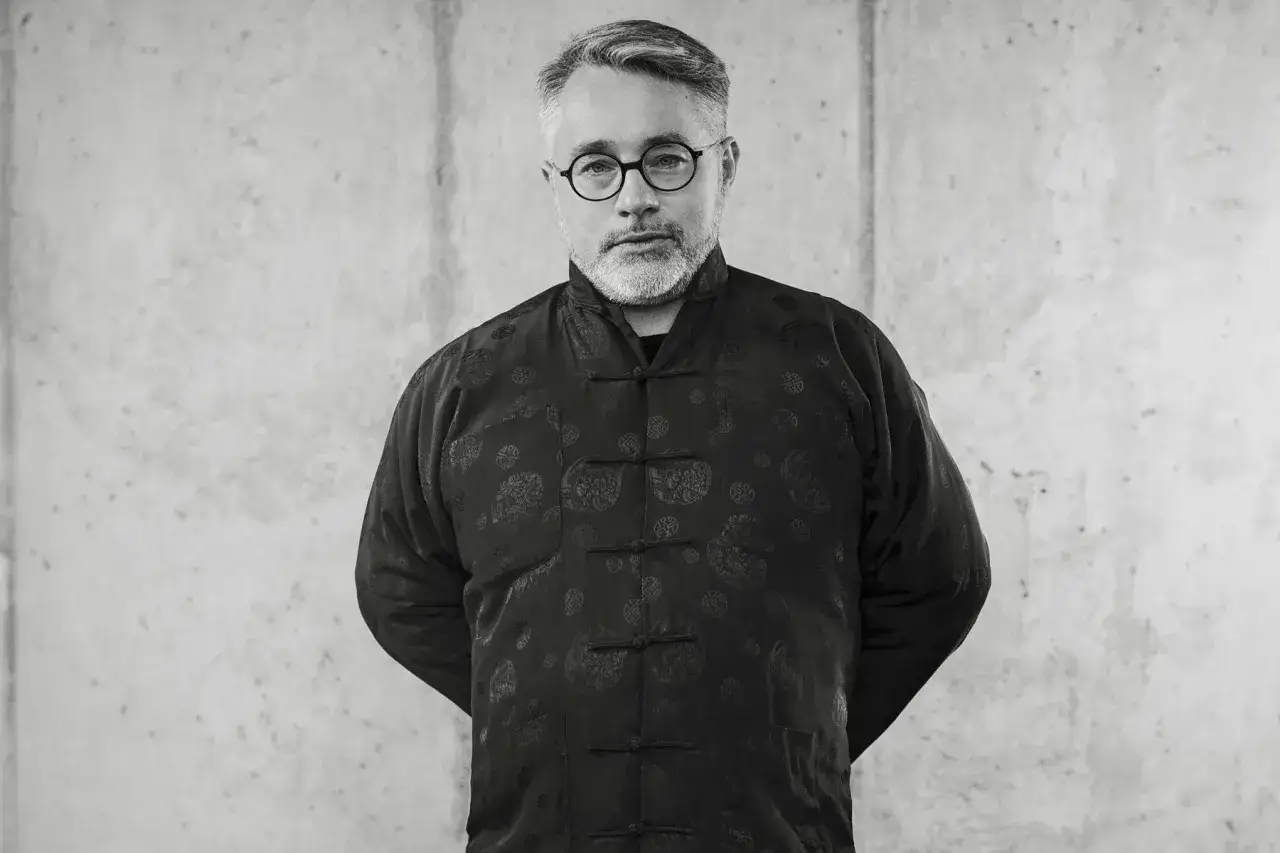 Mężczyzna w okularach i czarnym, wzorzystym robak wruhme stoi na tle betonu.