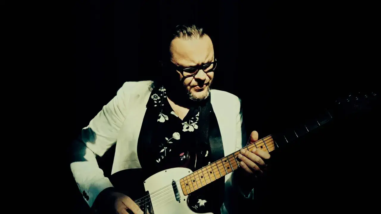 Mężczyzna w okularach i białej marynarce gra na gitarze. Jego styl przypomina zeppy zep.
