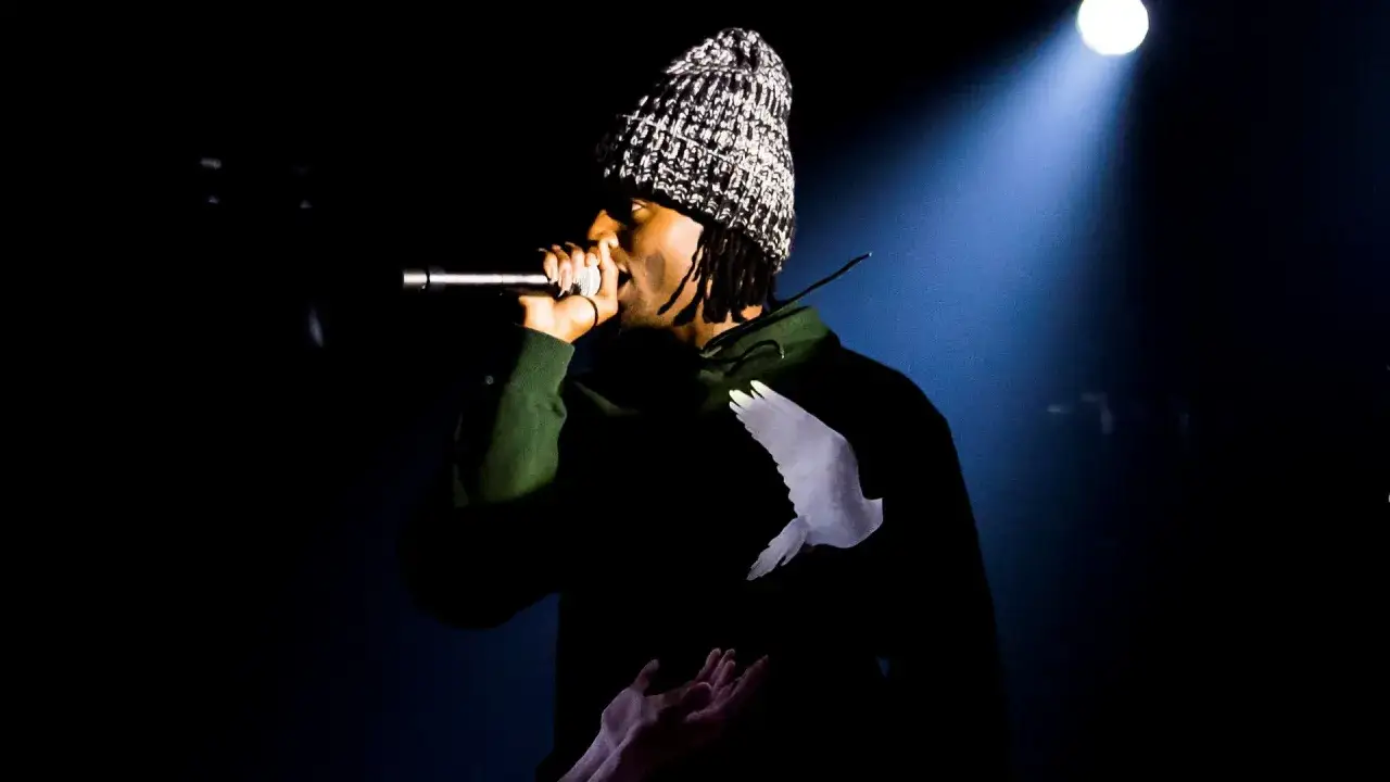 Playboi Carti na koncercie, w czapce i zielonej bluzie z gołębiem, śpiewa do mikrofonu.