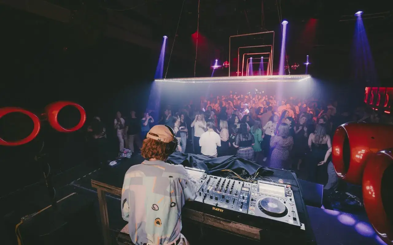 DJ techno w klubie, tłum tańczy w rytm muzyki, światła tworzą atmosferę.