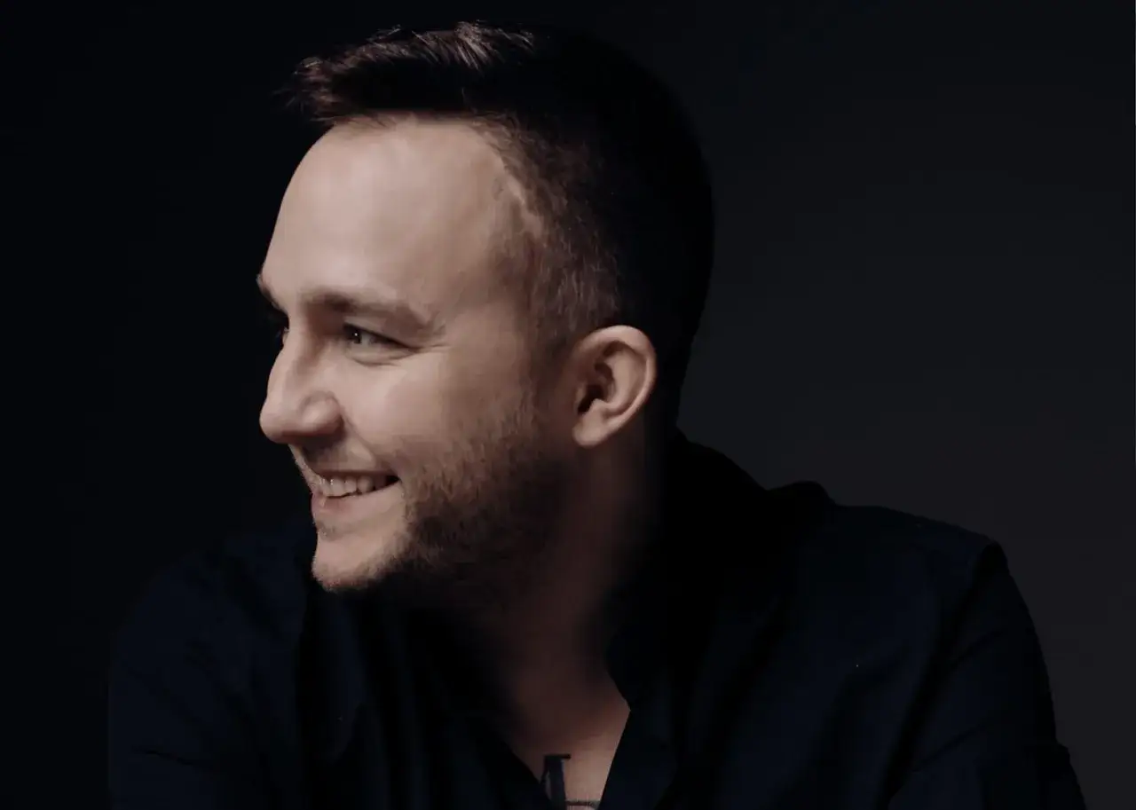 Uśmiechnięty mężczyzna w czarnej koszuli, w profilu. Ten typ mes wywiad, widać tatuaż na szyi.