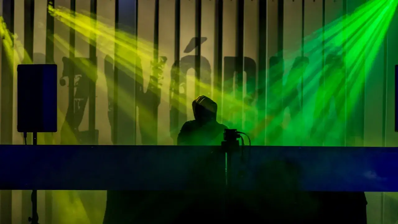 Artysta w kapturze na scenie, oświetlony zielonymi i żółtymi światłami. Tło z logo, sugerujące koncert tonfa rap.