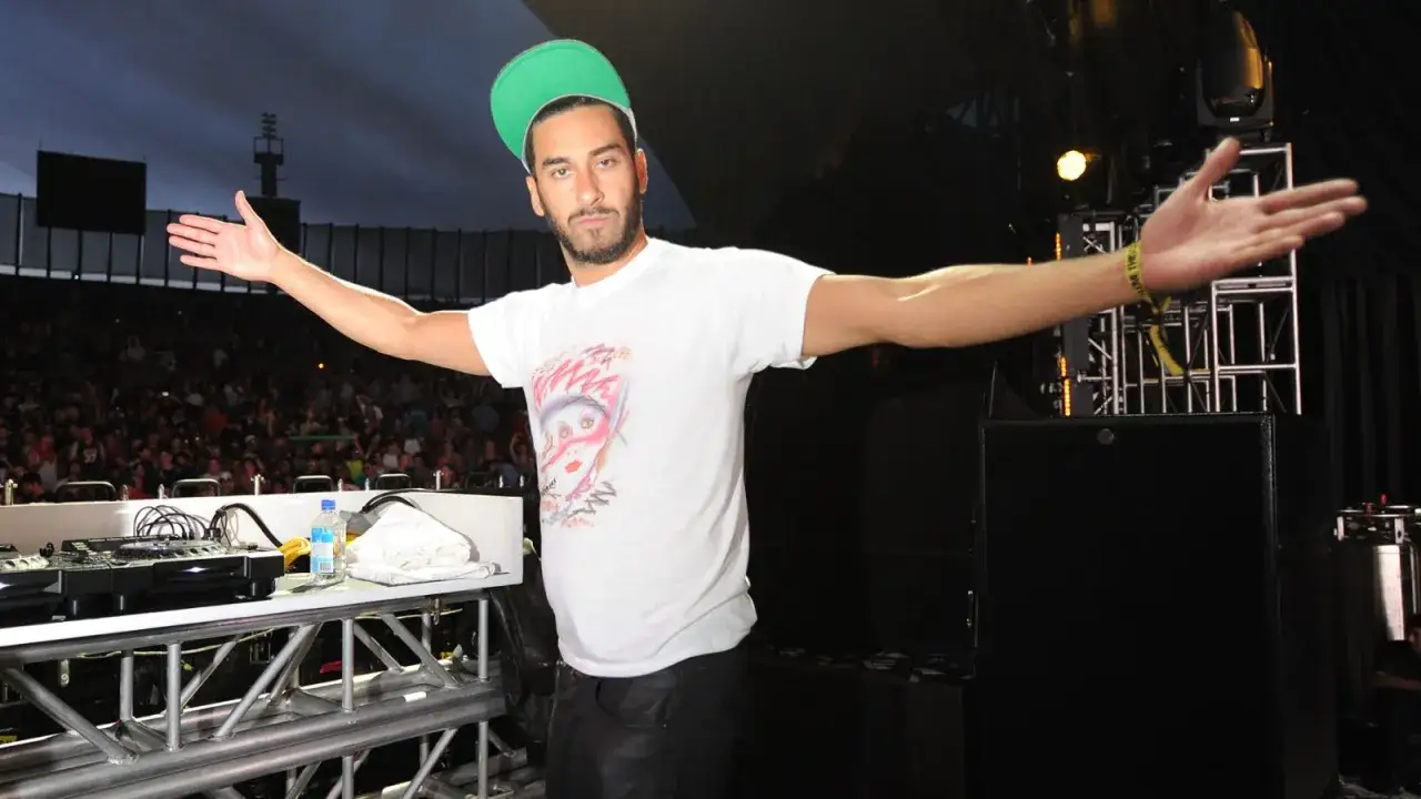 Armand Van Helden w zielonej czapce i białej koszulce z grafiką, z rękami rozłożonymi na tle tłumu i sceny.