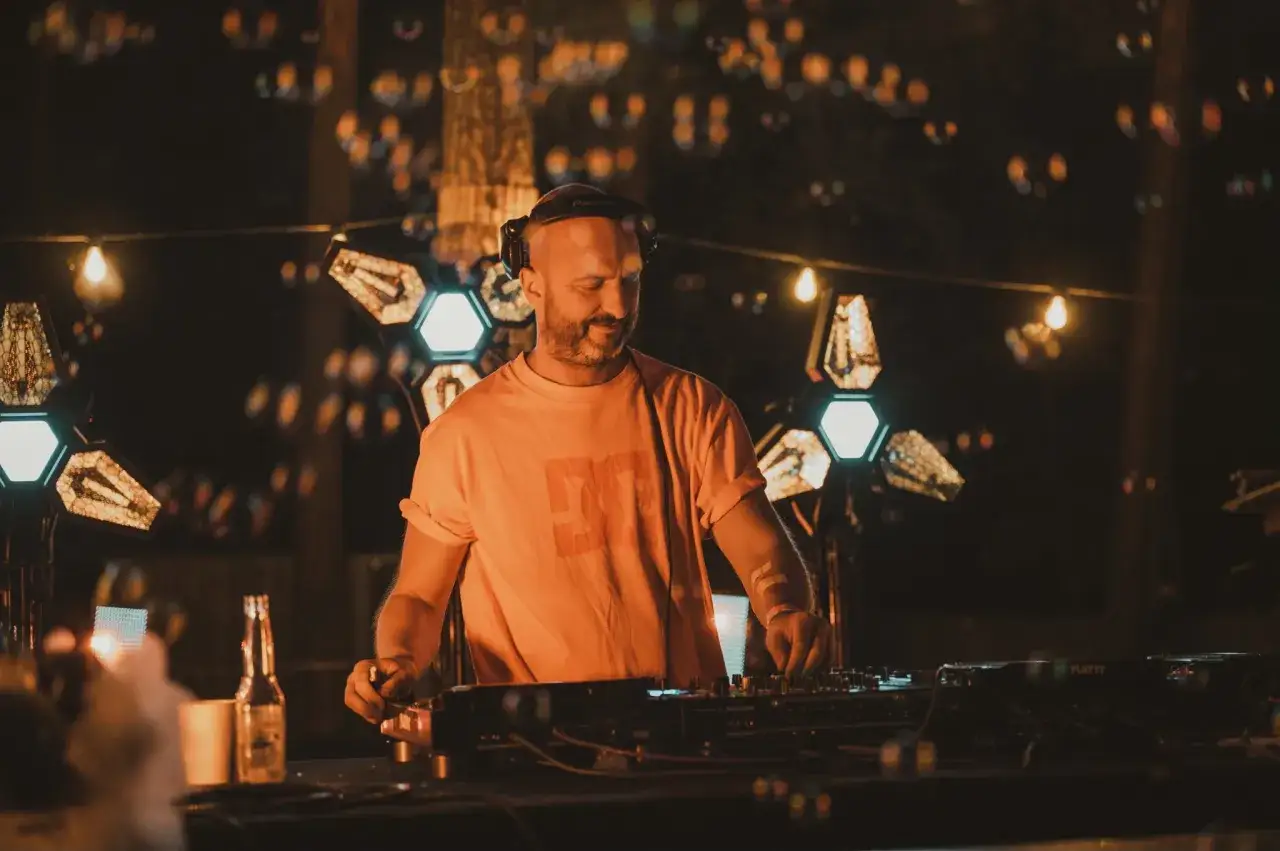 DJ Glasse w pomarańczowej koszulce miksuje muzykę na imprezie pod gołym niebem, otoczony światłami i bańkami mydlanymi.