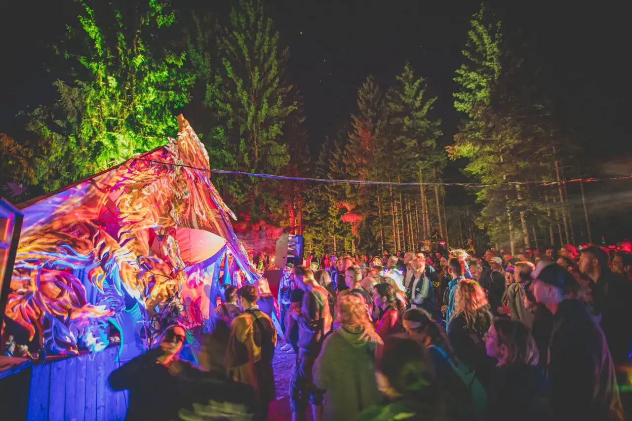 Tłumy ludzi bawią się na festiwalu techno pod kolorowo oświetlonymi drzewami.