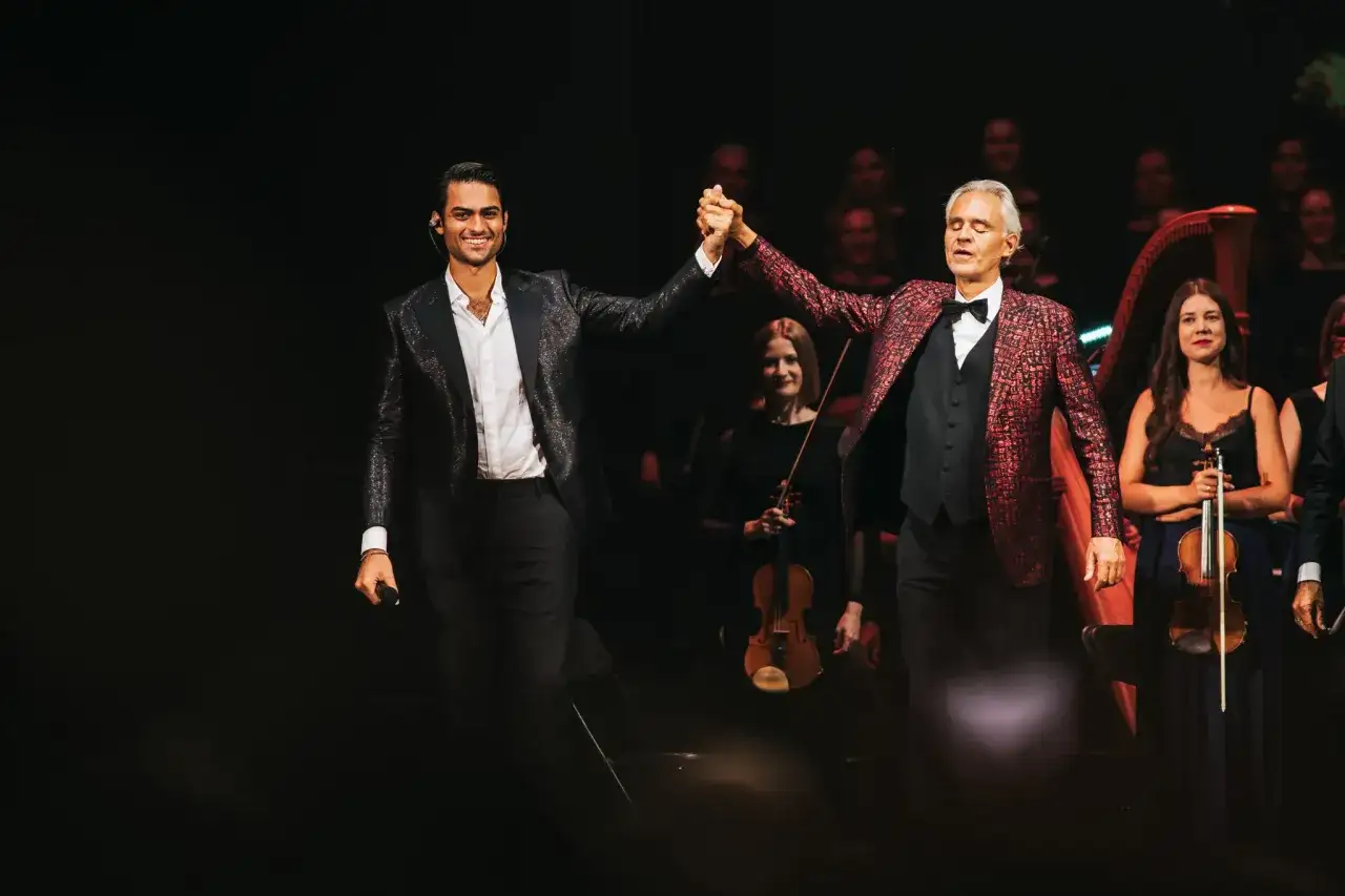 Dwóch artystów na scenie, jeden z mikrofonem, drugi z ręką w górze. W tle orkiestra i chór. Niezapomniany koncert Bocelli Warszawa.