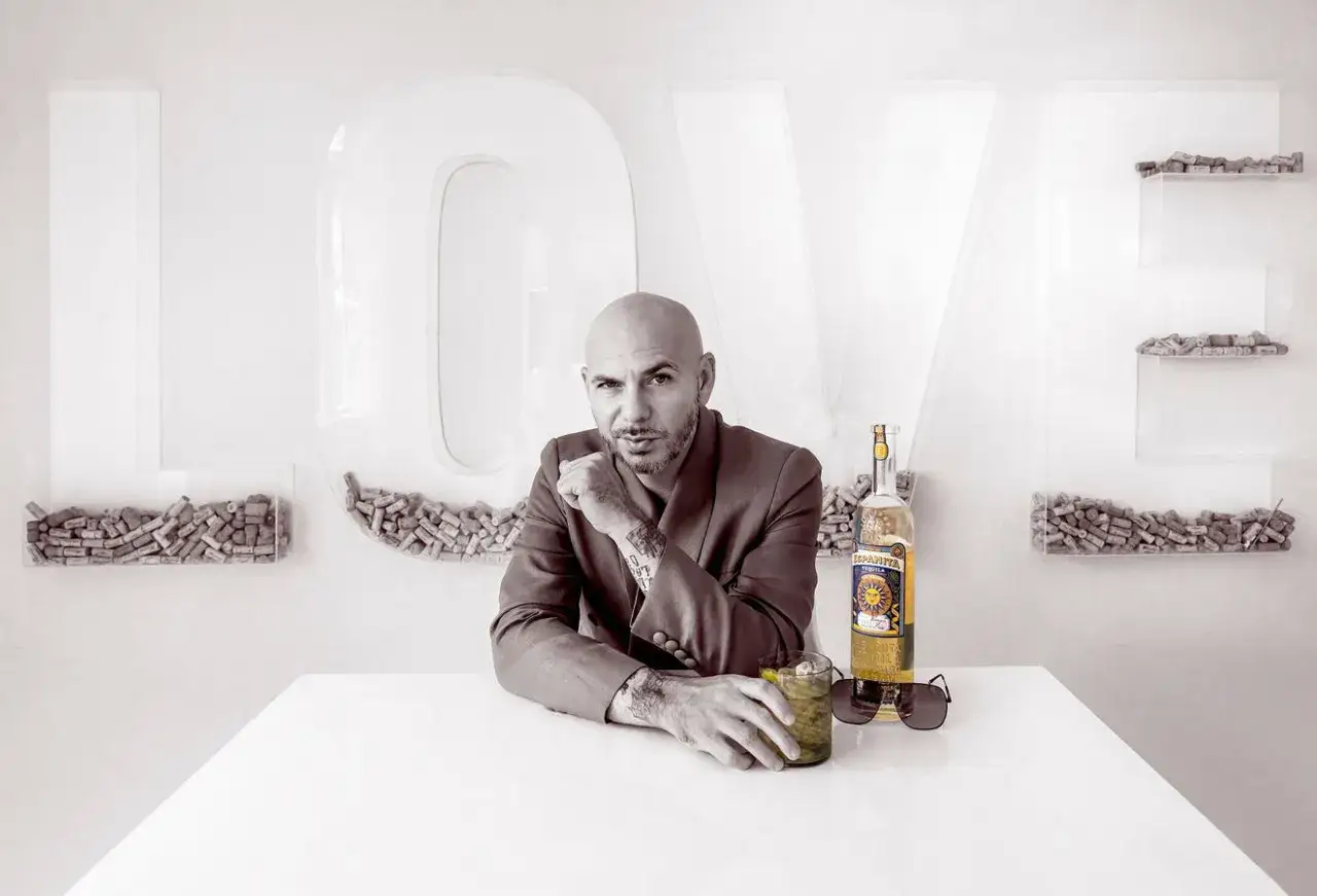 Pitbull, znany jako Mr Worldwide, siedzi przy stole z drinkiem i butelką tequili. W tle litery